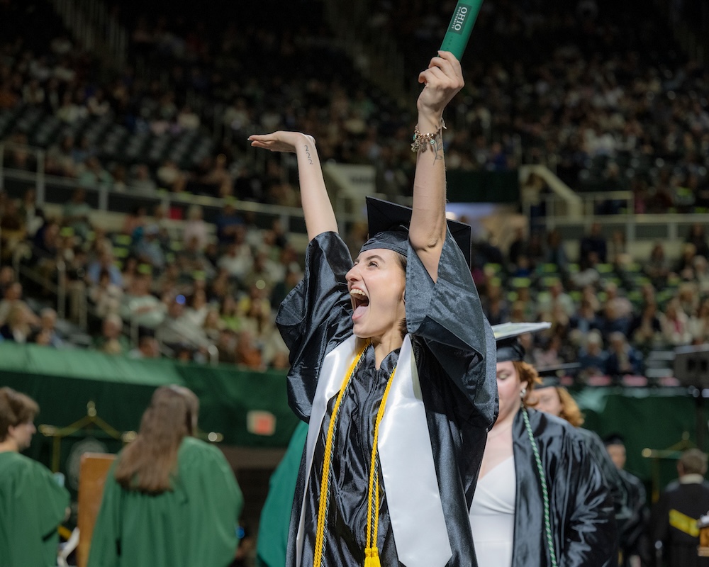 a student wearing a cap and gown raises her hands in triumph holding a new diploma