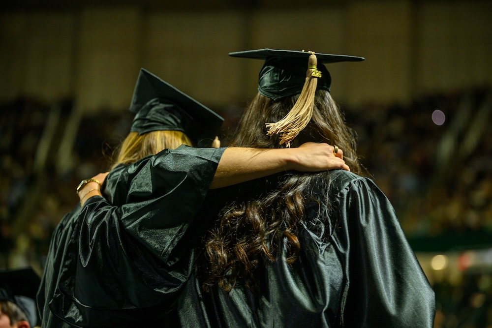 Pictured from behind, two OHIO students stand with their arms around each other wearing caps and gowns at the 2026 Commencement ceremony