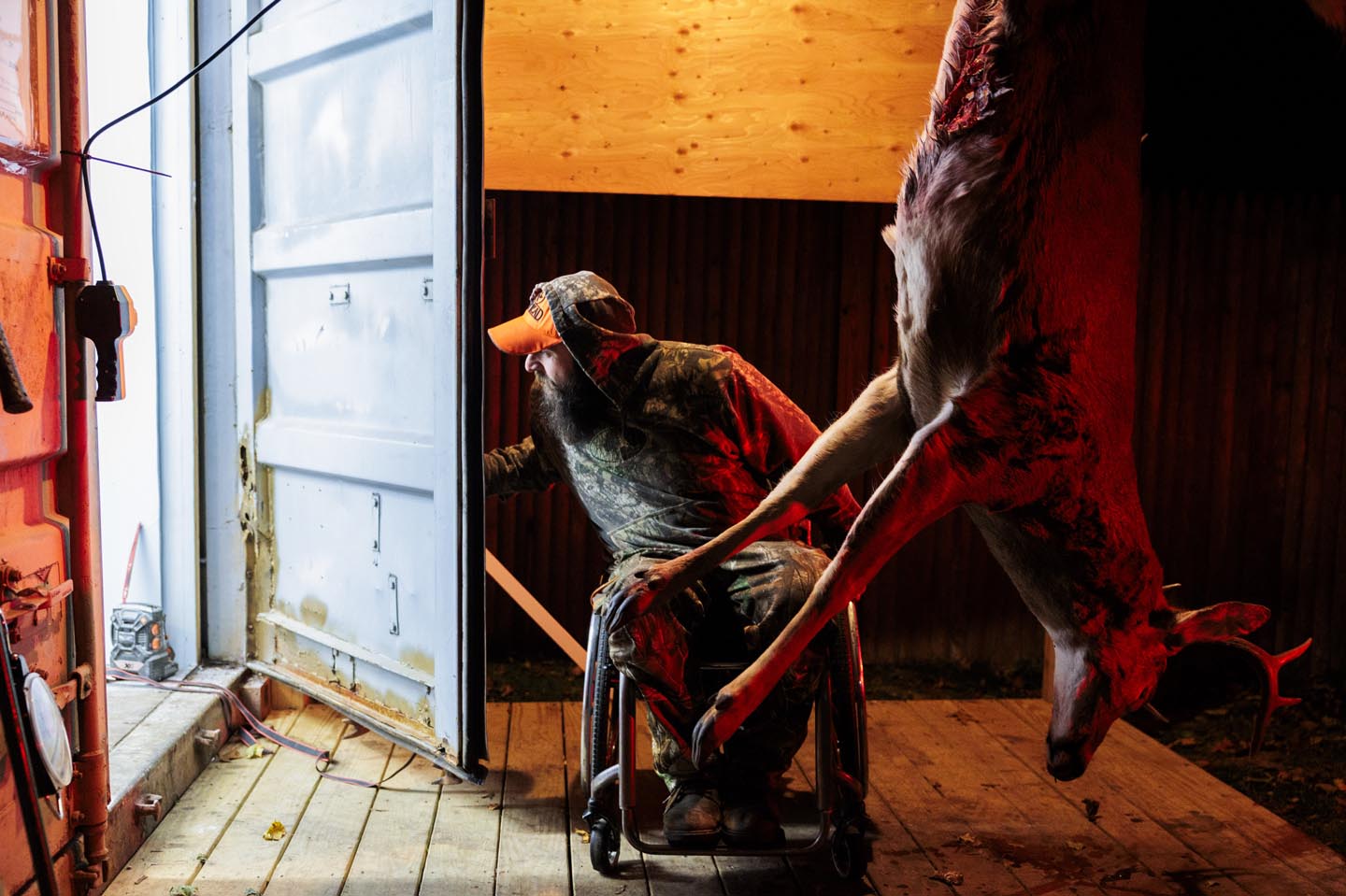 A man in a wheelchair looks out a doorway; a deer carcass hangs in the foreground