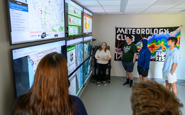 OHIO students gather around weather screens as part of a meteorology class. A sign for a meteorolgy club is also shown in the image.