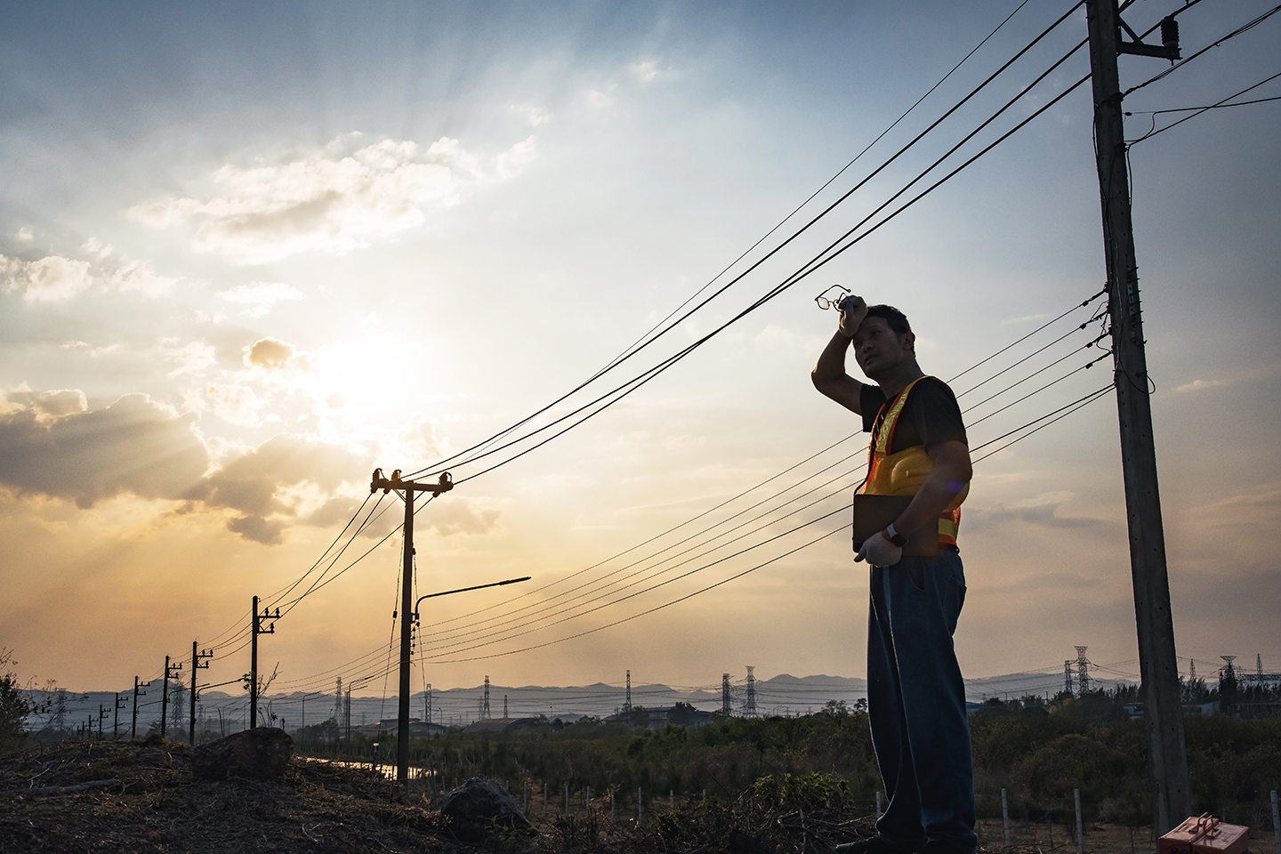 A power line worker wipes sweat off of his brow while standing in front of power lines.