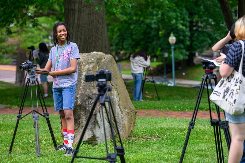 A camp participant smiles at other campers while also taking video on the College Green. Several other participants are also recording video nearby.