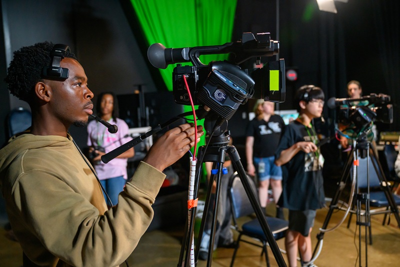 A camp participant records video through a large camera while other participants are also recording video and working on projects inside an OHIO building.