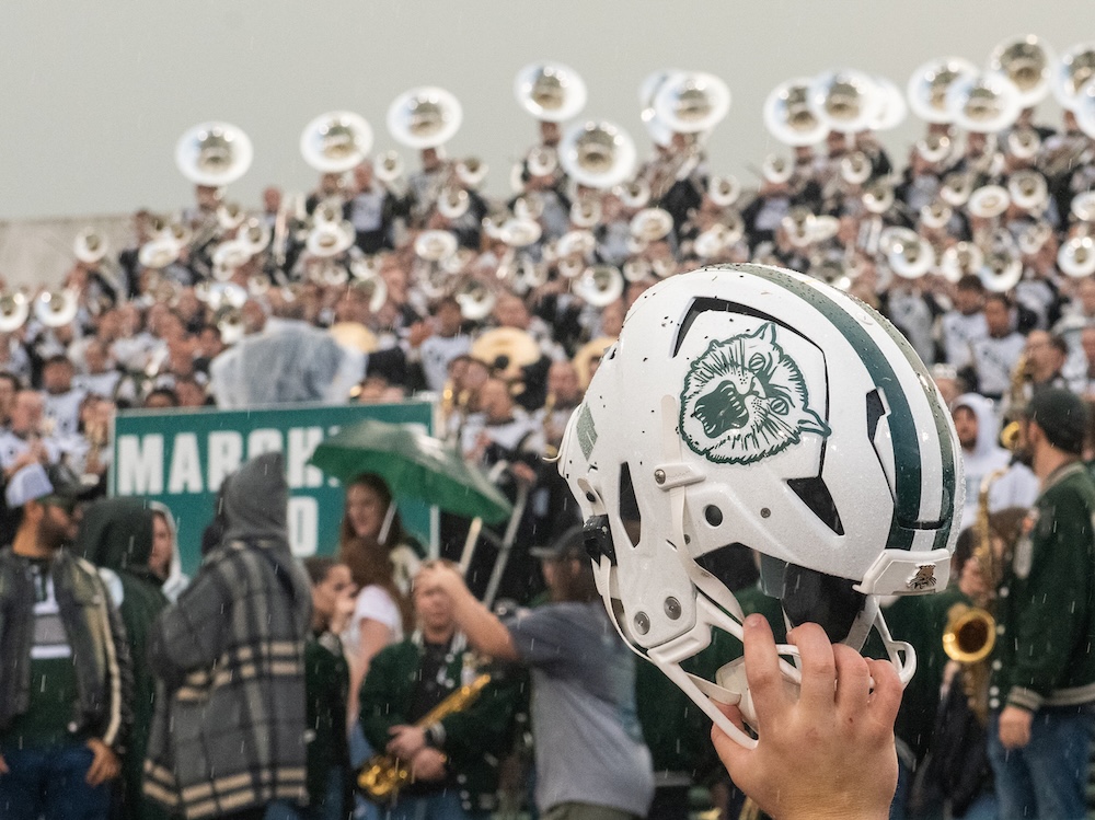 An OHIO football helmet pictured in the foreground of a rainy stadium, with the horns of the marching band in the background