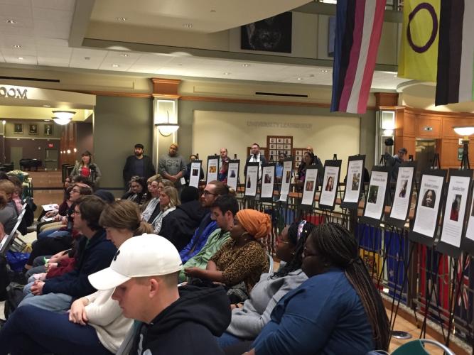 The crowd at the Trans* Day of Remembrance ceremony sits in front of posters of victims of violence in the U.S.