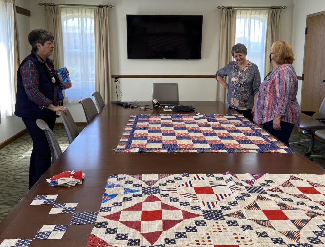 Three women stand around a board table, with two unfinished quilts resting on the table