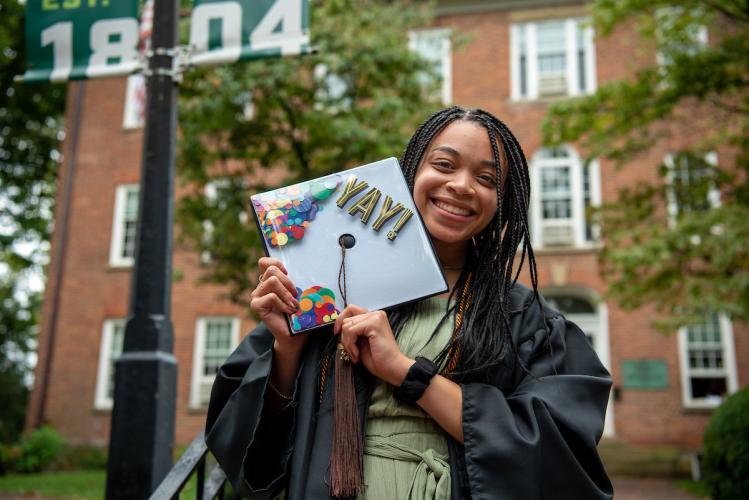 A graduate poses with her cap that reads "YAY!" with artwork to look like confetti.
