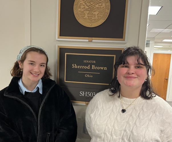 Two college students pose for a picture standing on either side of a sign that says "Senator Sherrod Brown"