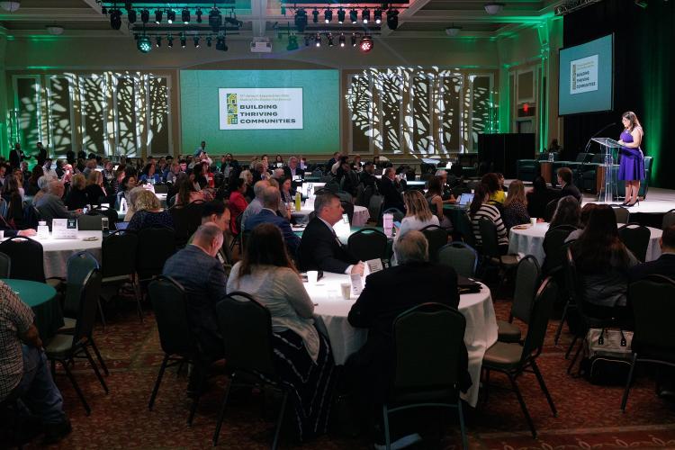 A speaker talks to a large crowd seated at tables in the Baker Center Ballroom for the State of the Region Conference - A slide on the wall states Building Thriving Communties