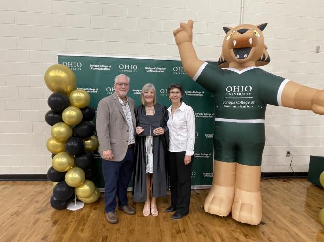 Mary Stephan is shown in her commencement robe, holding her cap, standing with Scott Titsworth and Ann Bainbridge while they stand next to balloons and a blow-up figure of Rufus wearing a Scripps College of Communication shirt