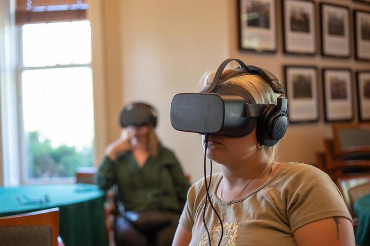 Two individuals wear virtual reality headsets while sitting at tables at Ohio University.