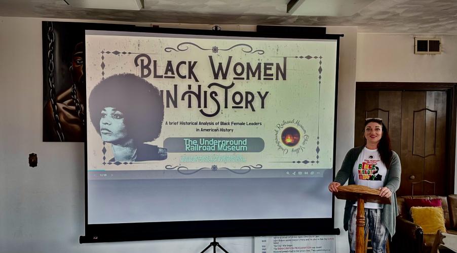 Photo of a female with long black hair standing behind a wooden podium, next to a projection screen that show a slide stating "Black Women in History"