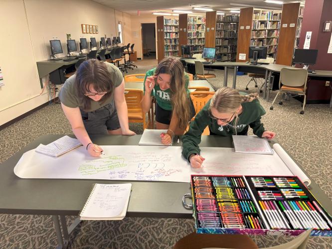 Three students use markers on large posterboard
