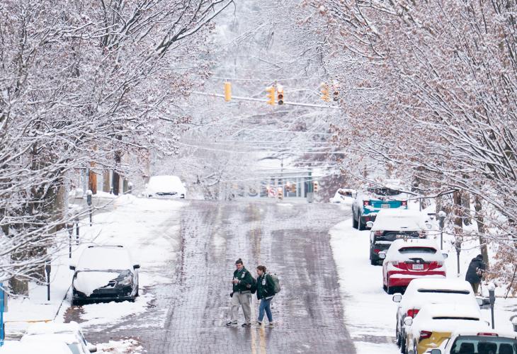 Two OHIO students walk across a street while the cars near them and the trees above them are covered in snow