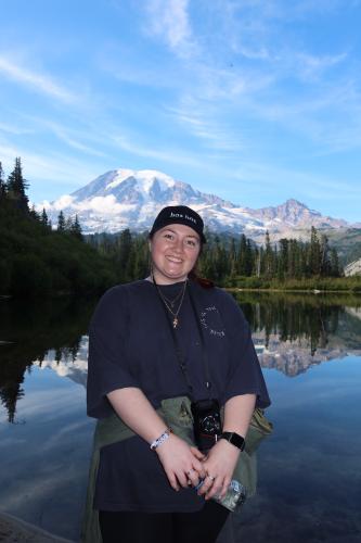 Jenna Pfeiffer stands in front of a large body of water with a mountain, tall trees and a blue sky in the background.