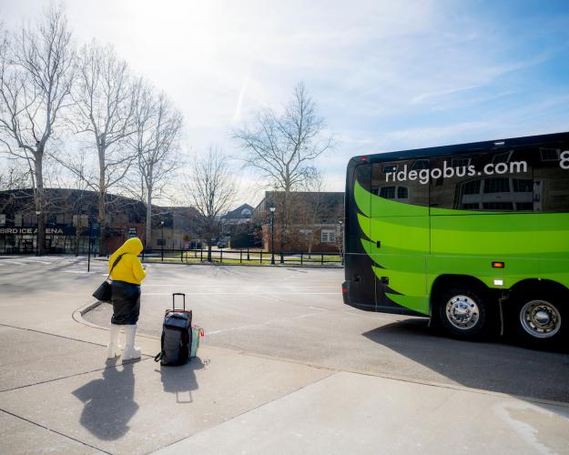 An OHIO student with a suitcase  waits outside of a GoBus parked near Baker University Center, Bird Arena and the Aquatic Center
