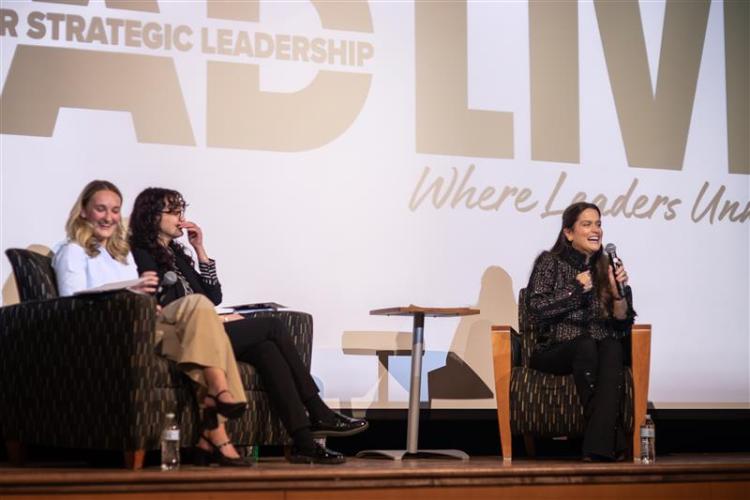 Louisa Vilela speaks while sitting in a chair on the stage at the Lead Live event, while Grace Cantwell and Kasielle Carroll sit in chairs nearby on the stage.