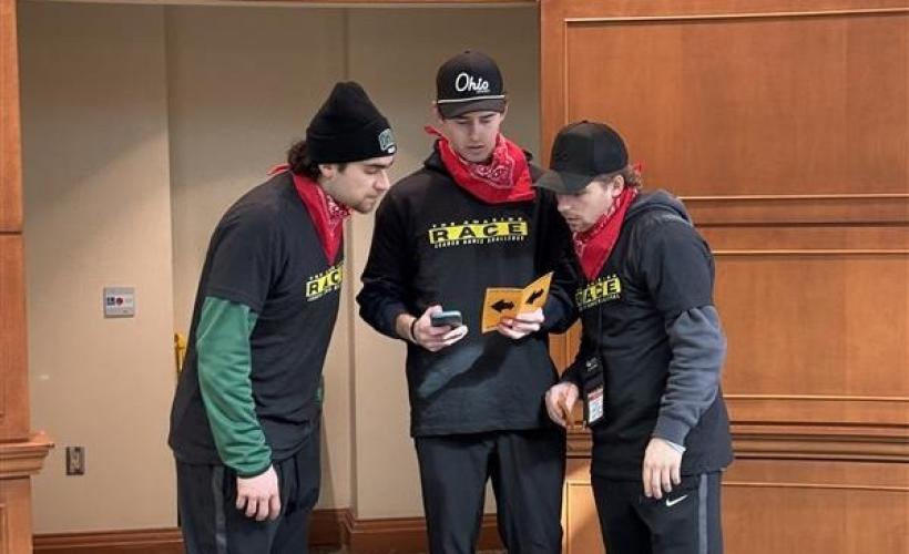 Three participants in the Leader Games Challenge wear Amazing Race shirts and look over information about the game while standing in Walter Rotunda
