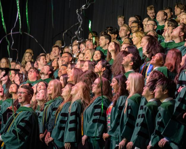 A large group of Heritage College graduates in green doctoral regalia look up and smile as green and silver streamers fall over them during their commencement ceremony.