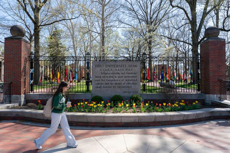 An OHIO student walks in front of the College  Gate, which has colorful flowers in front of it and colorful trees behind it, on a spring day