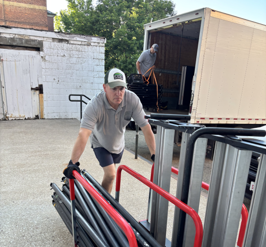 One member of the Moving and Surplus Team moves a cart filled with poles and other items outside while another member of the team moves items in the back of a moving truck