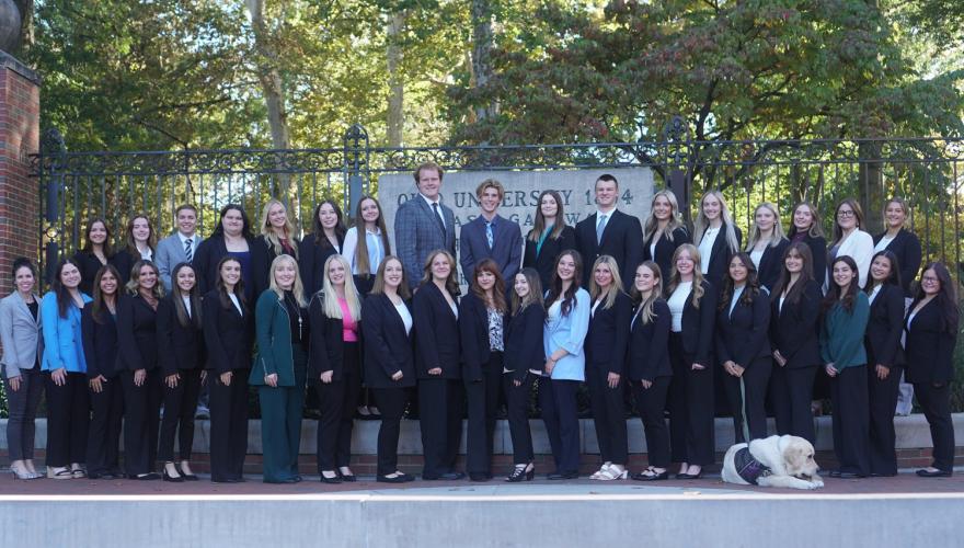 OHIO students dressed in professional attire stand in two rows in front of the College Gate, a dog sits at the feet of one student