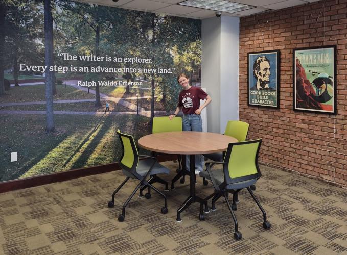 An OHIO student stands by a round table at the renovated Writing Commons. The table is in front  of a wall that has an image of the College Green and the words "The writer is an explorer. Every step is an advance into a new land - Ralph Waldo Emerson"