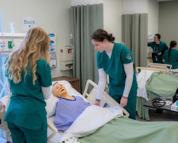 Students in scrubs attend to manikin in hospital bed
