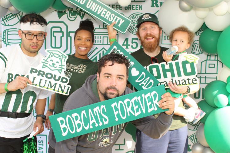 Four adults and a child pose in front of a OHIO-themed backdrop, featuring green colors and prideful signs at the Homecoming Family Tent.