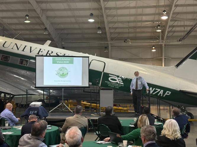 A speaker gives a presentation in an airplane hangar while standing on a stage in front of an Ohio University plane