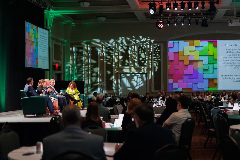 Five panelists talk on stage in front of a very large crowd in the Baker Center Ballroom
