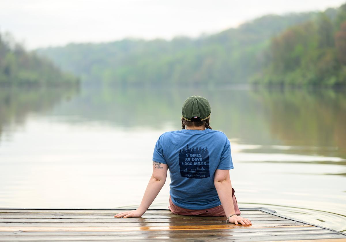 Helena Karlstrom sits on a dock at Strouds Run State Park while wearing a shirt that says "4 Girls, 85 Days, 1,200 miles"