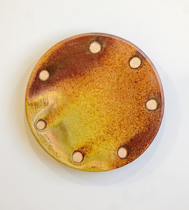 a circular ceramic piece in shades of orange and yellow, with seven lighter orange circles spaced semi-evenly around its circumference 