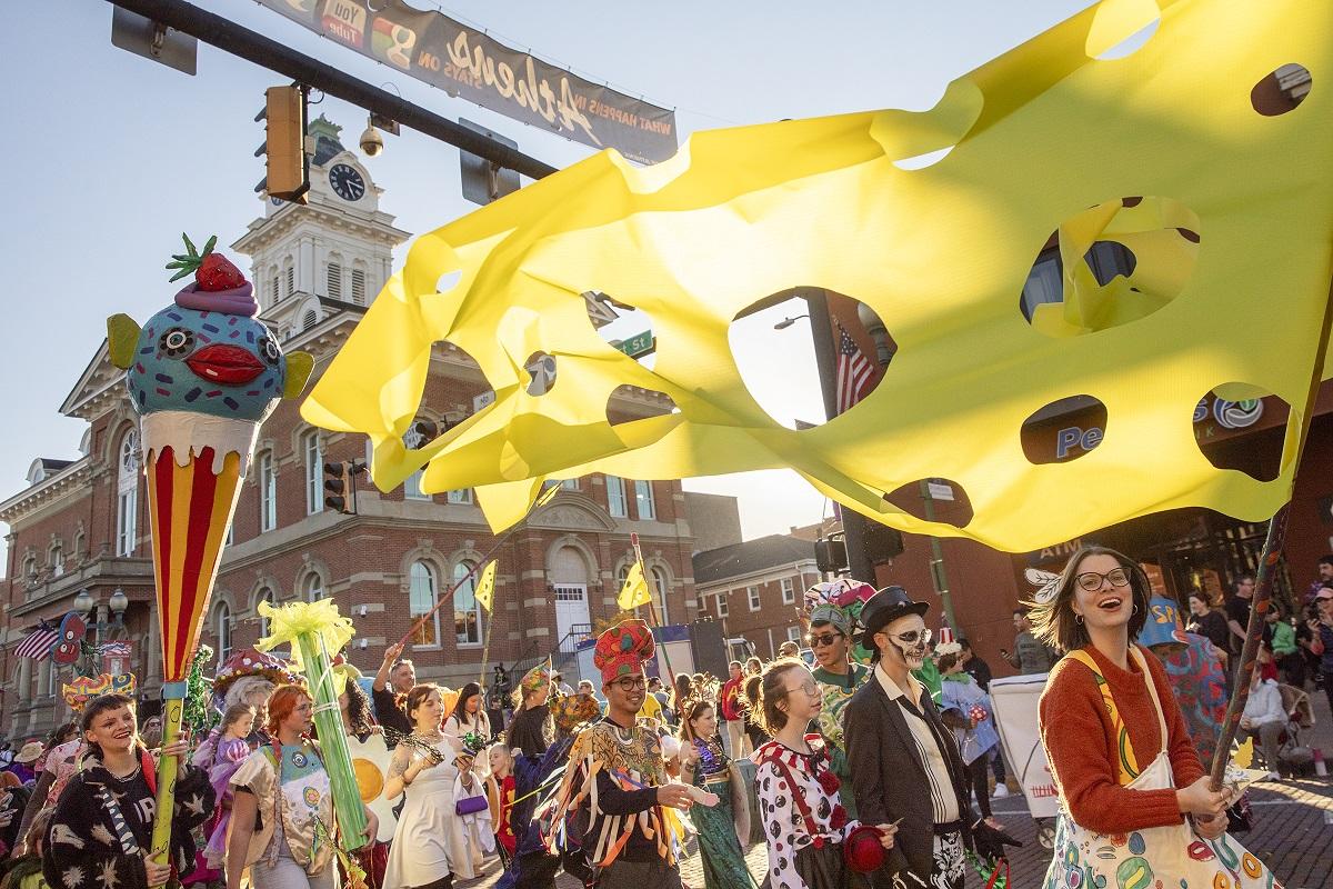 People in costumes march in the Honey for the Heart parade in Athens while one person carries a flag that looks like a giant piece of cheese