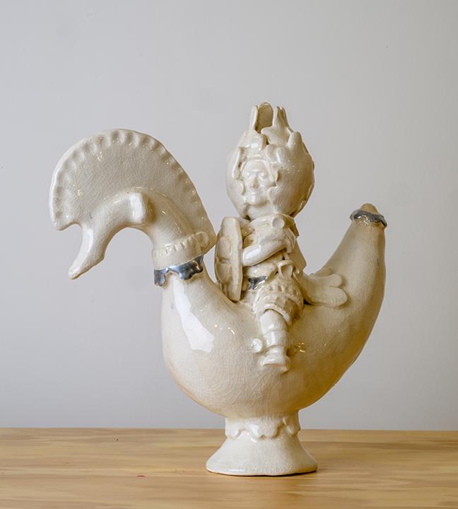 a white ceramic statue of a person wearing a helmet and carrying a shield, sitting astride a birdlike figure
