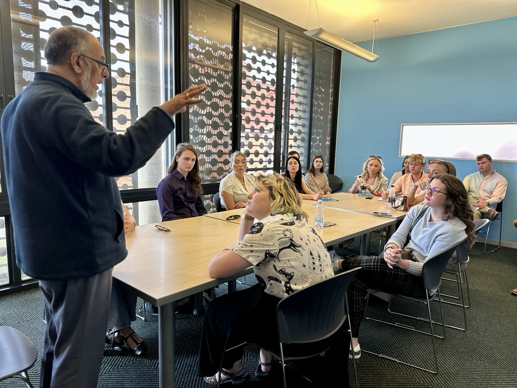 An OHIO faculty member speaks in front of a group of students who are sitting around a table.