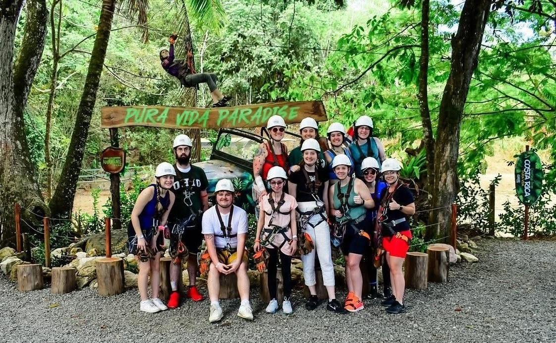 Ohio University students are shown wearing zip line gear as they stand together at a zip line park in Costa Rica, and a sign behind them says "Pura Vida Paradise"
