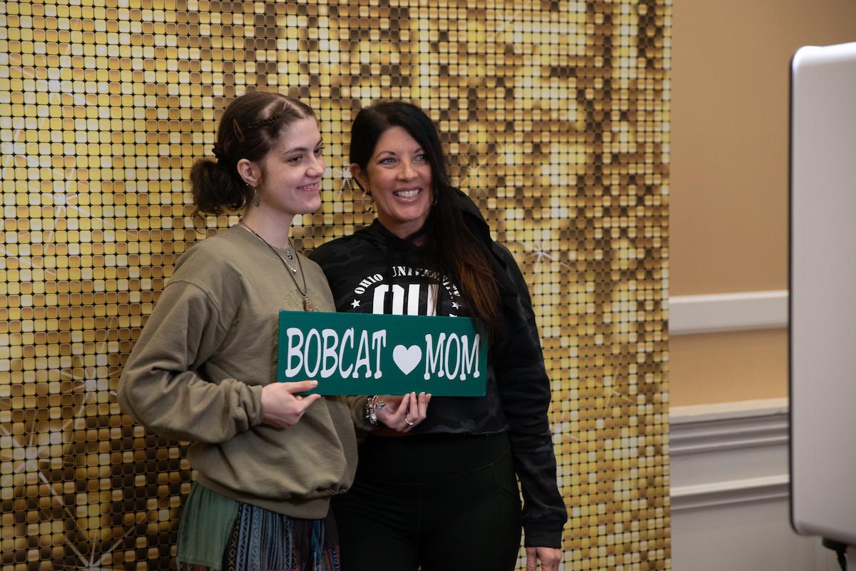 An OHIO student and a parent stand together with a sign that says Bobcat (heart) Mom