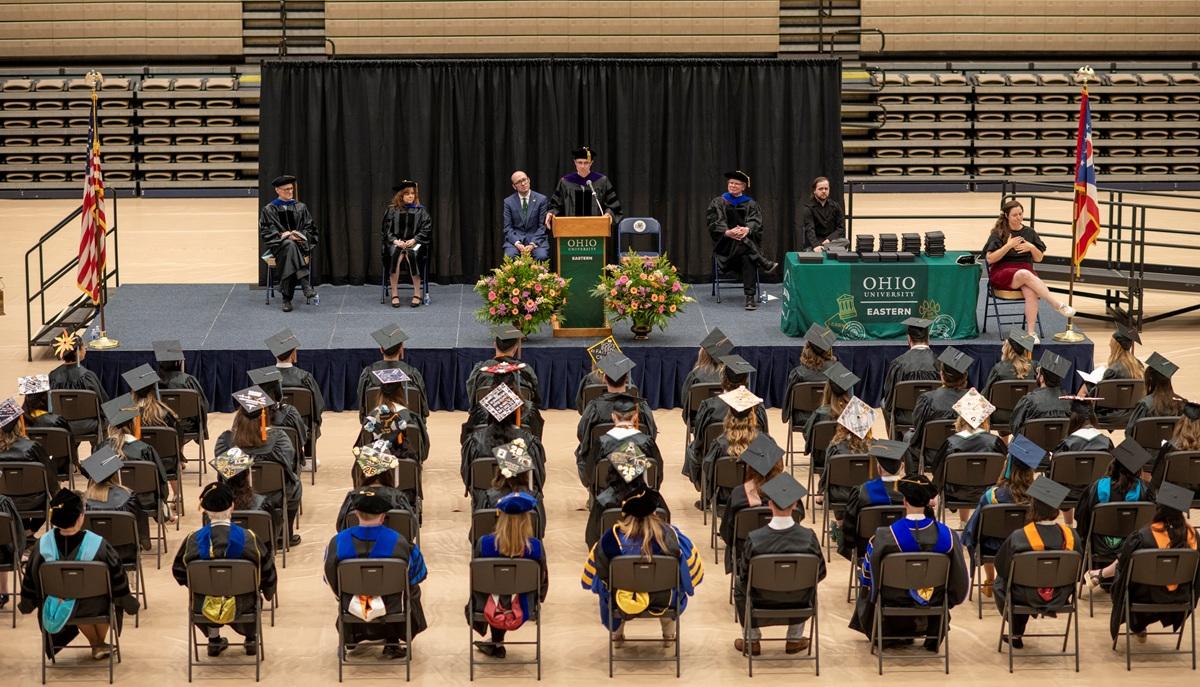 OHIO graduates sit in rows of chairs in a gymnasium while they wear caps and gowns and listen to a speaker at the podium during the Graduation Recognition Ceremony at Ohio University Eastern. 