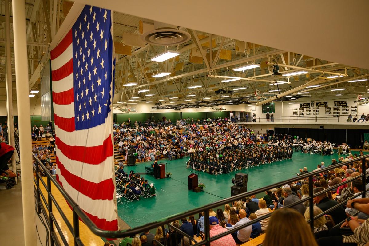 The graduates sit togeher in the middle of the floor while they are surrounded by a large crowd in the gymnasium  for the Graduation Recognition Ceremony at Ohio University Chillicothe. A large USA flag hangs at the top of the gym, too.