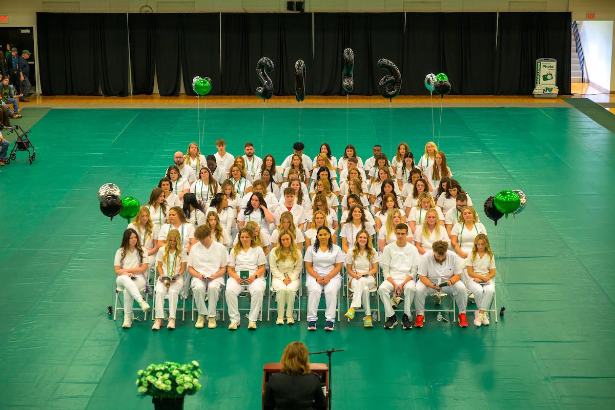 OHIO Chillicothe Nursing students sit in chairs on the floor in the gymnasium while a speaker stands at the podium during the Pinning Ceremony
