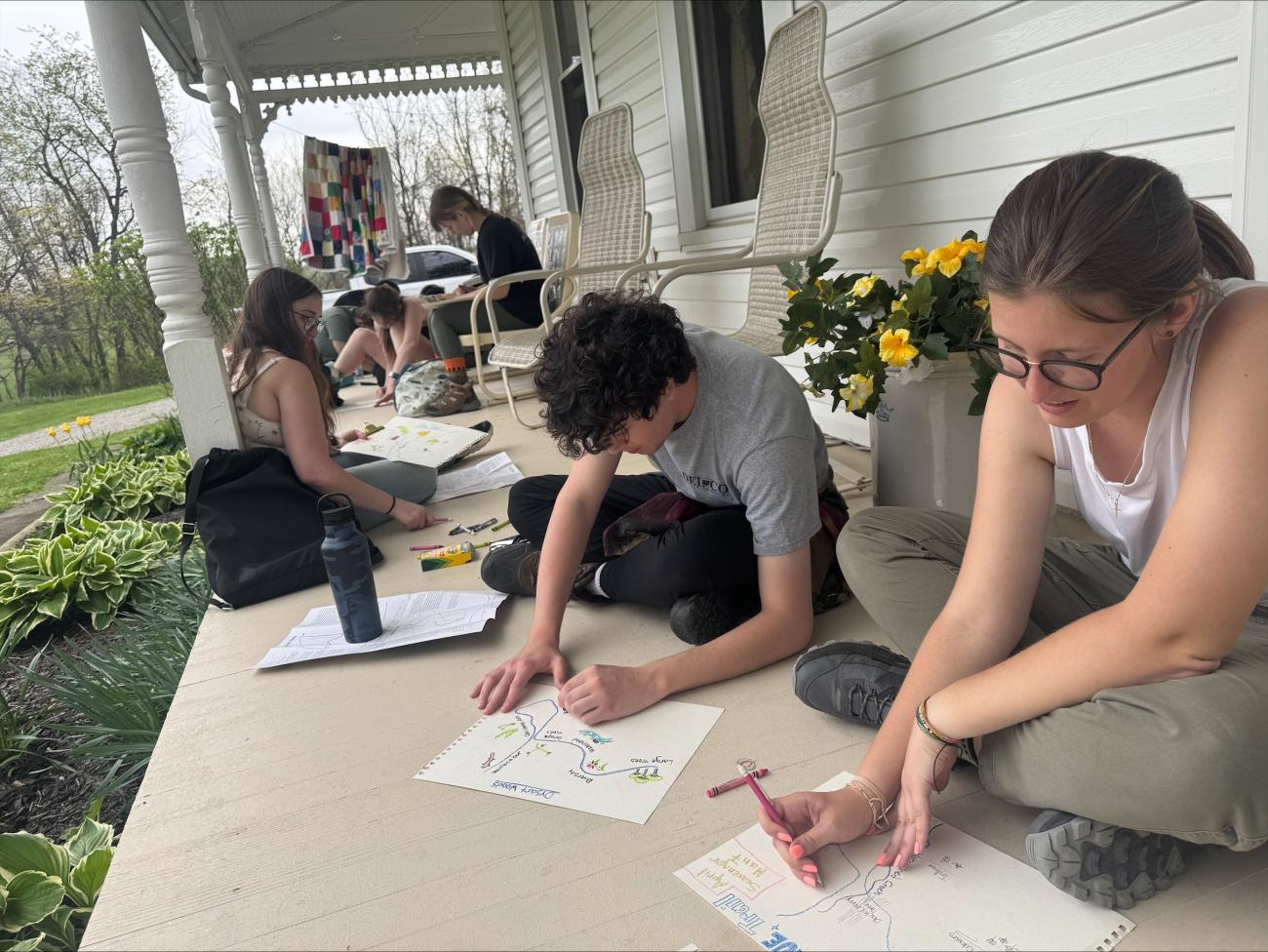 Students sitting on a concrete porch, drawing maps on white paper.