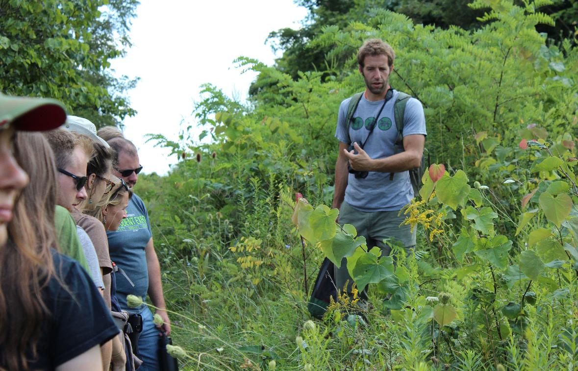 Joe Brehm is shown talking in tall grass and brush in the woods on a summer day, while a group of people stands nearby.