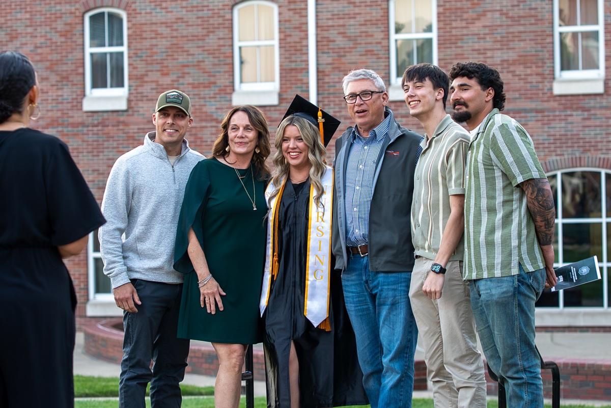 An OHIO graduate wearing the graduation cap and gown stands with family members in front of a large brick building after the Graduation Recognition Ceremony at Ohio University Southern.