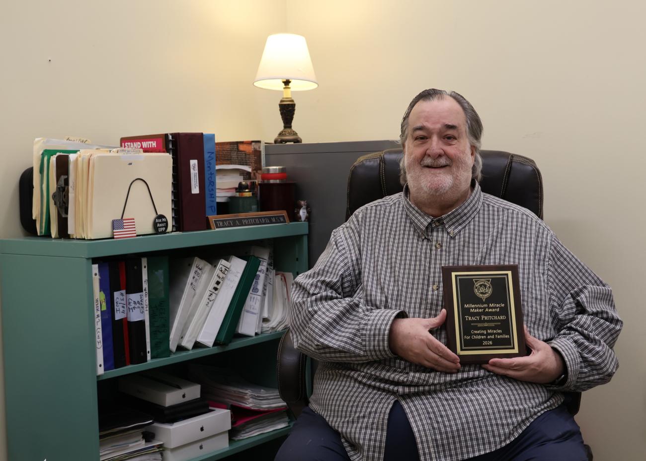 Tracy A. Pritchard holding an award, seated in a chair in his office in front of a teal bookcase filled with books and files