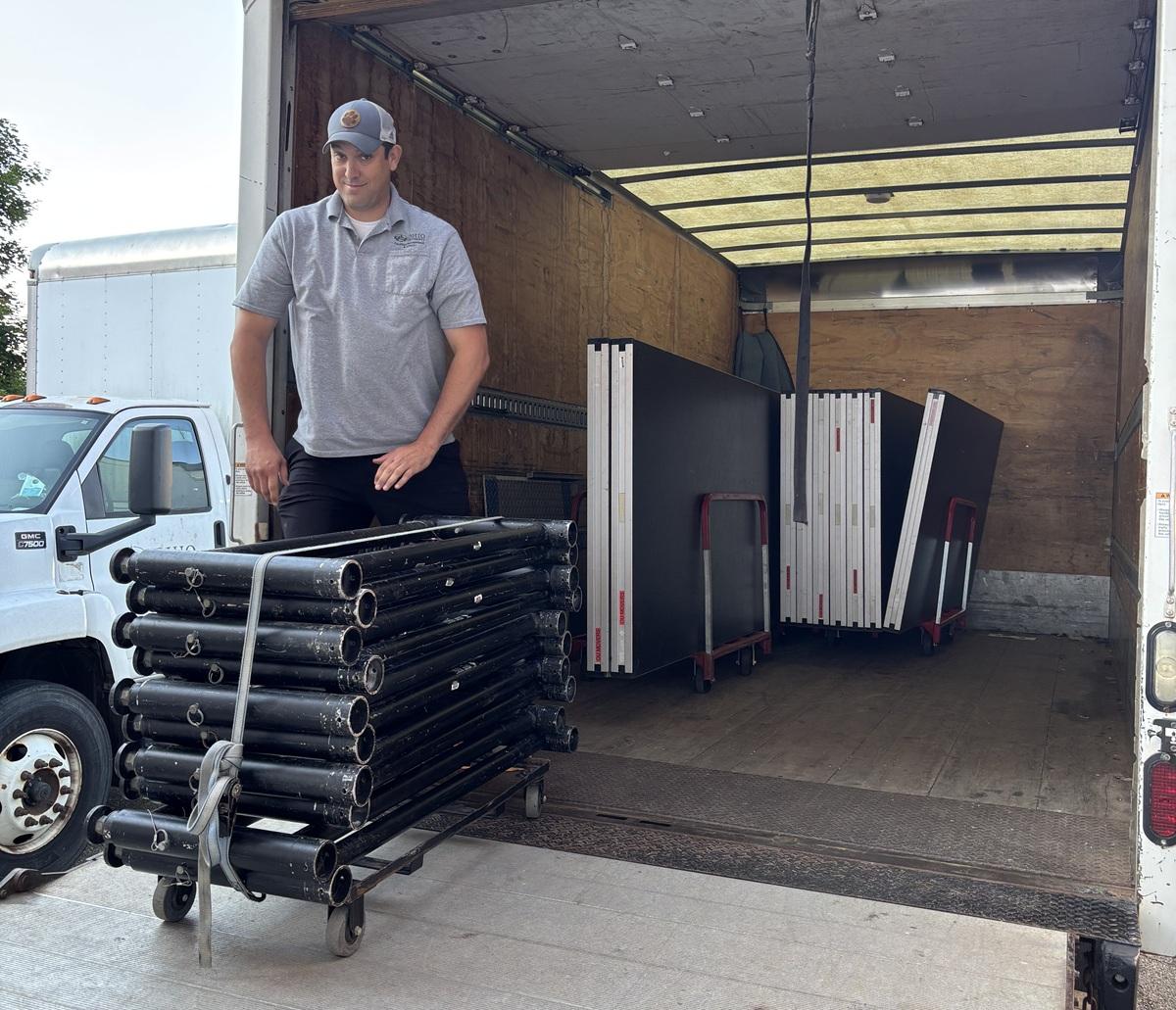 A member of the Moving and Surplus Team stands in the back of a moving truck next to  a cart filled  with items, and in front of moving carts filled with large items
