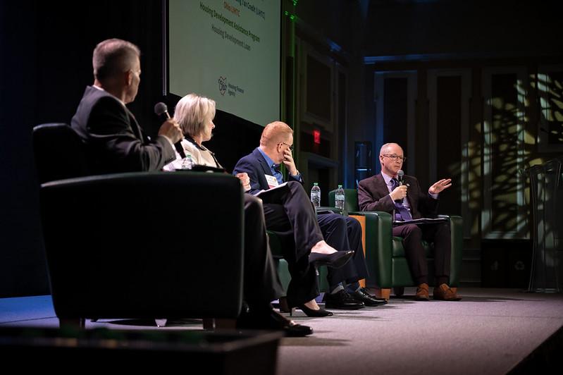 Four panel members sit on the stage and talk during a panel discussion event at the State of the Region Conference