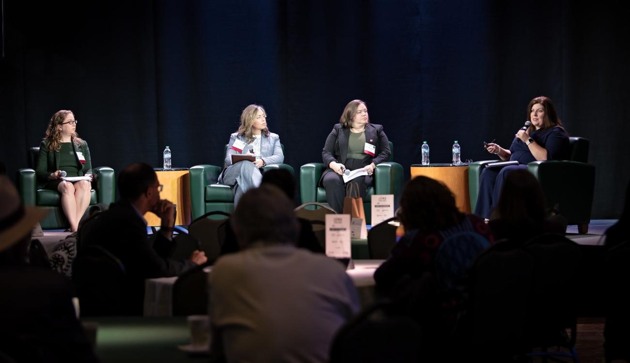 Four panel members sit on the stagee, while one of them talks into a microphone during a panel discussion event at the State of the Region Conference