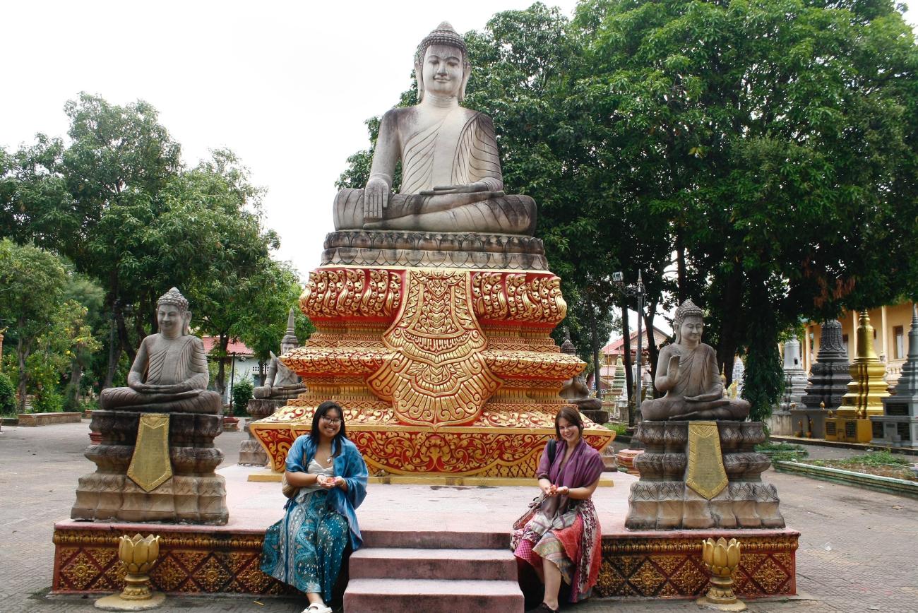 Andrea Tiu and Calli Fout sit in front of a large sculpture while in Cambodia