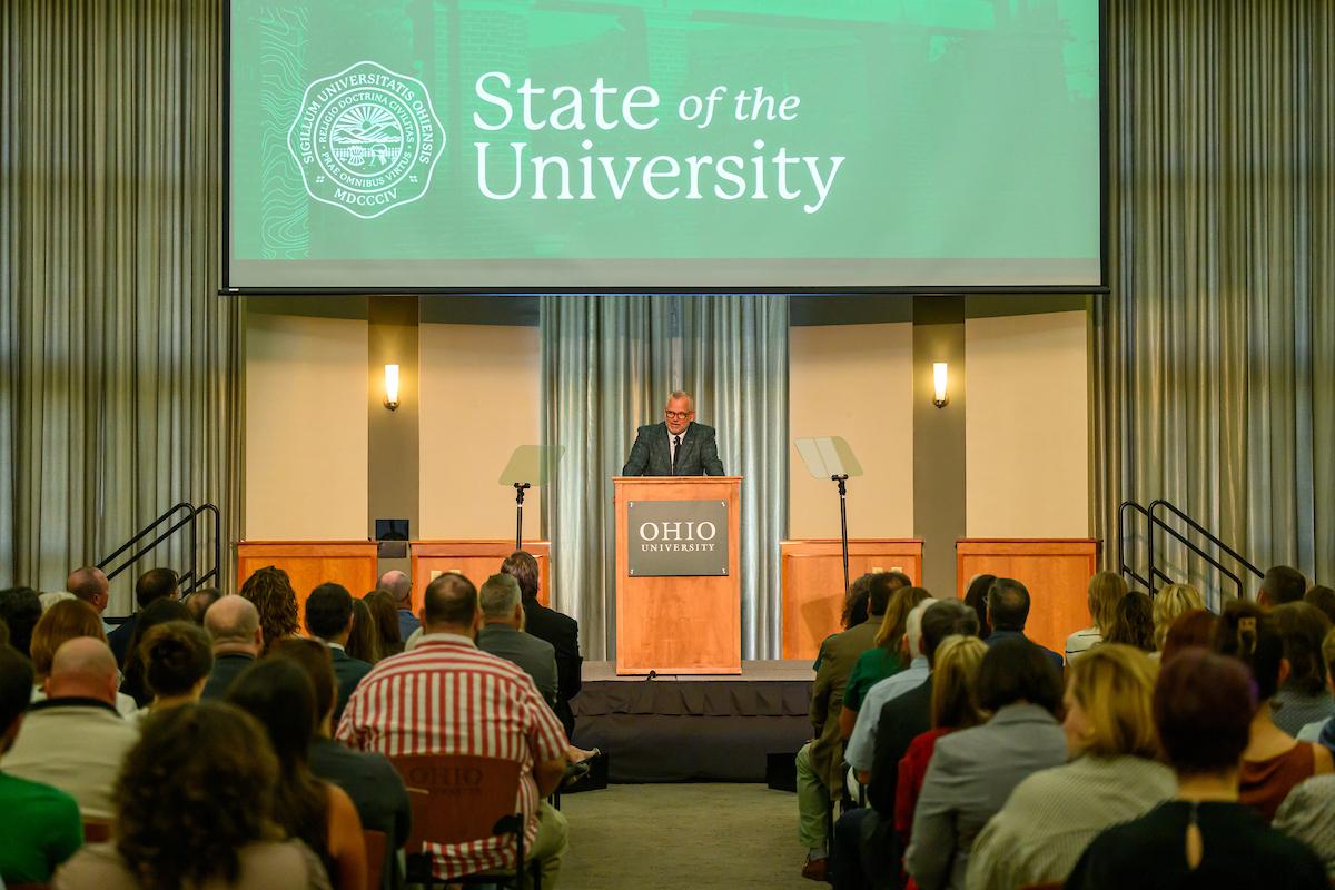 Matt Evans speaks at the podium in front  of a large crowd in Walter Hall, while the words State of the University are shown on the screen above him